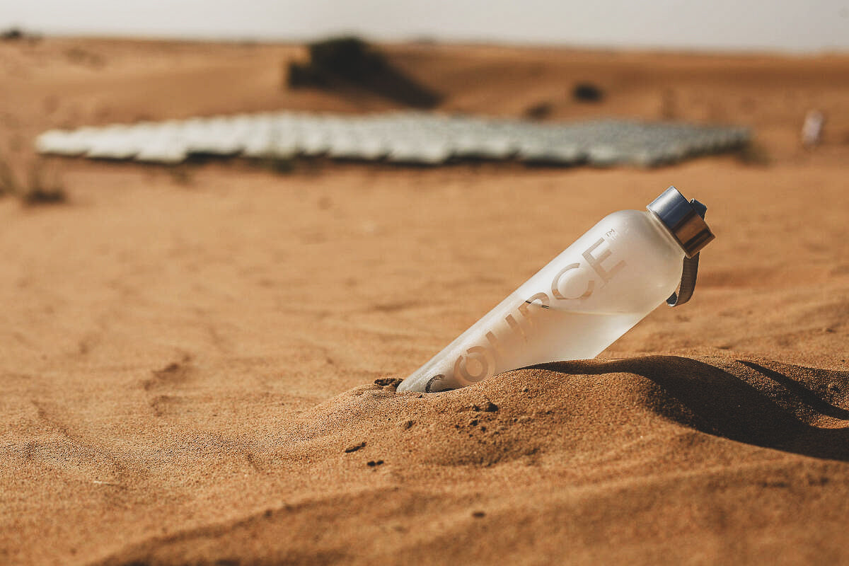 SOURCE reusable water bottle with renewable water in it sits in the sand with SOURCE panels in the background